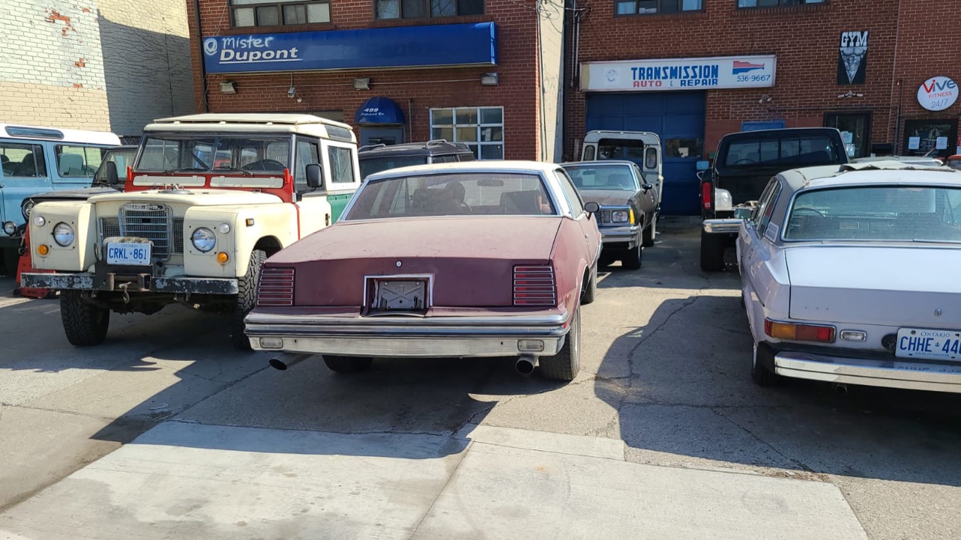 Grand Prix parked between two Land Rover Series trucks and a sedan outside a transmission shop